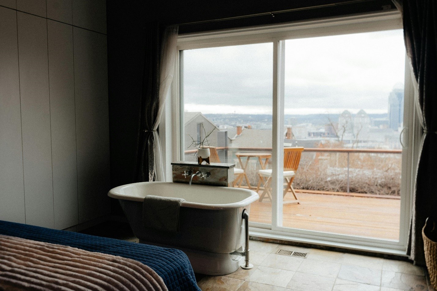 image of a bath tub in a bedroom which faces out onto a balcony and a city skyline