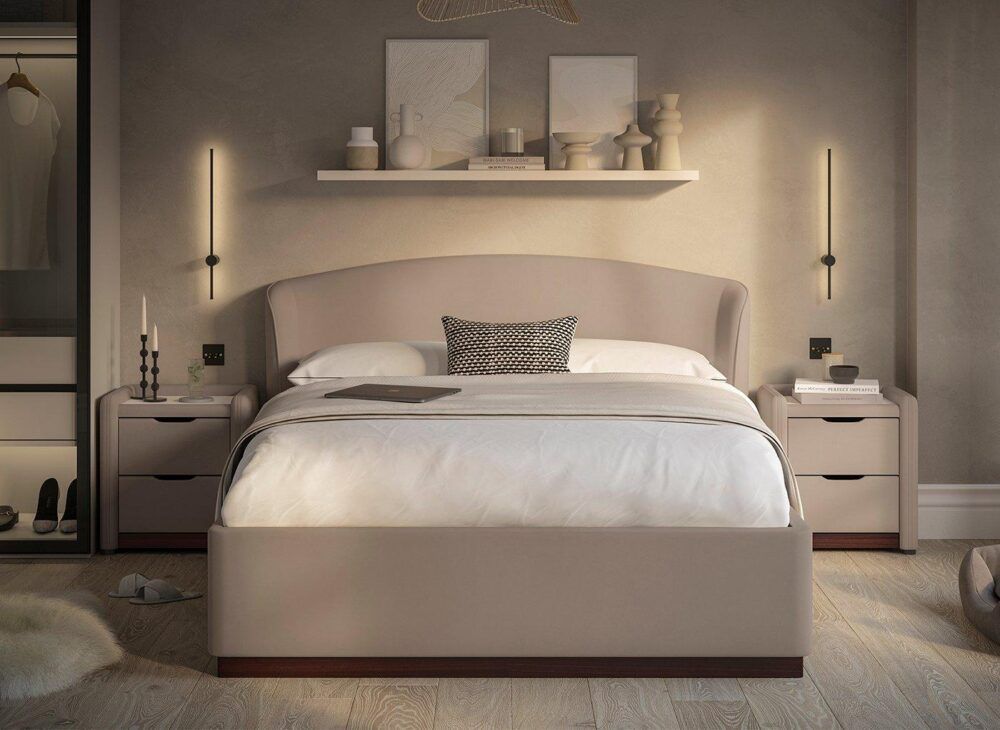 a taupe bed frame viewed from the foot end. The bed is placed centrally with a floating shelf above holding picture frames, vases, and ornaments.