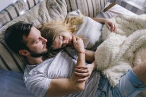 A couple cuddle together in bed, dressed in day clothes and with blankets.