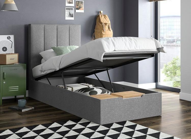 a bedroom with a grey ottoman single bed with the mattress lifted to show the storage. There's a geometric rug beneath one corner of the bed and covering a dark wooden floor. To the left of the bed is a metal green bedside table with a speaker on top and record player artwork above.