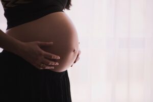 A pregnant person holds their tummy with both hands against a white curtain.