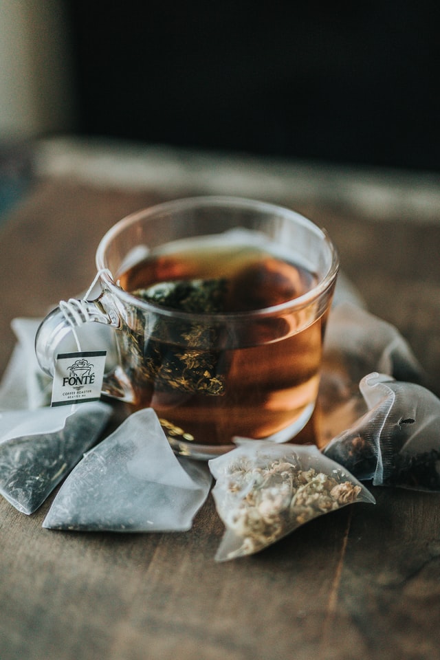 A brewing cup of tea in a glass, surrounded by different teabags on a wooden table.