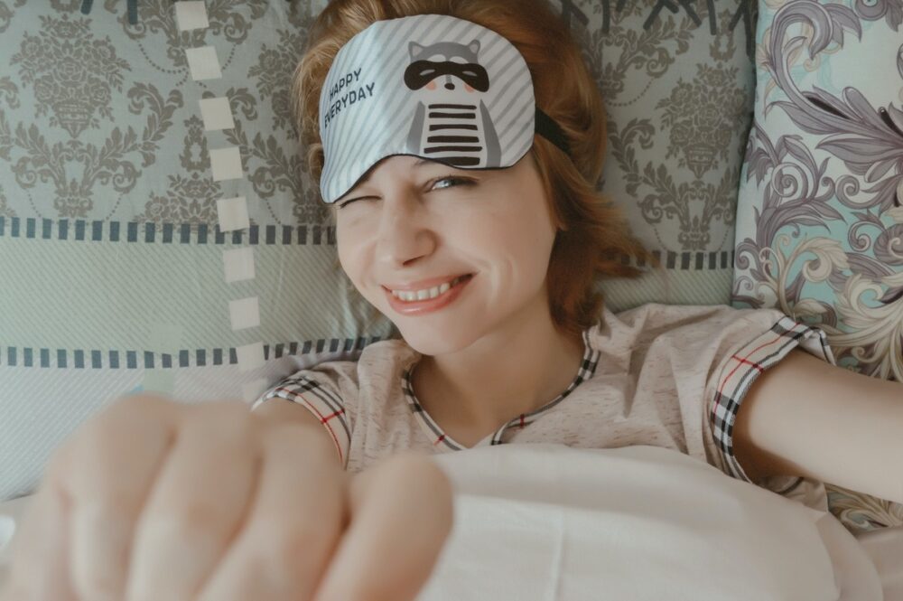 A lady wakes up in bed, smiling, with a sleep mask off her face.