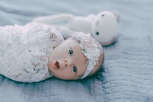 A baby swaddled in a white blanket, with a white headband, on a grey bed next to a white soft toy