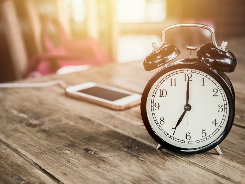 A phone sits on a wooden table, plugged in to charge. Next to it is an old fashioned alarm clock.